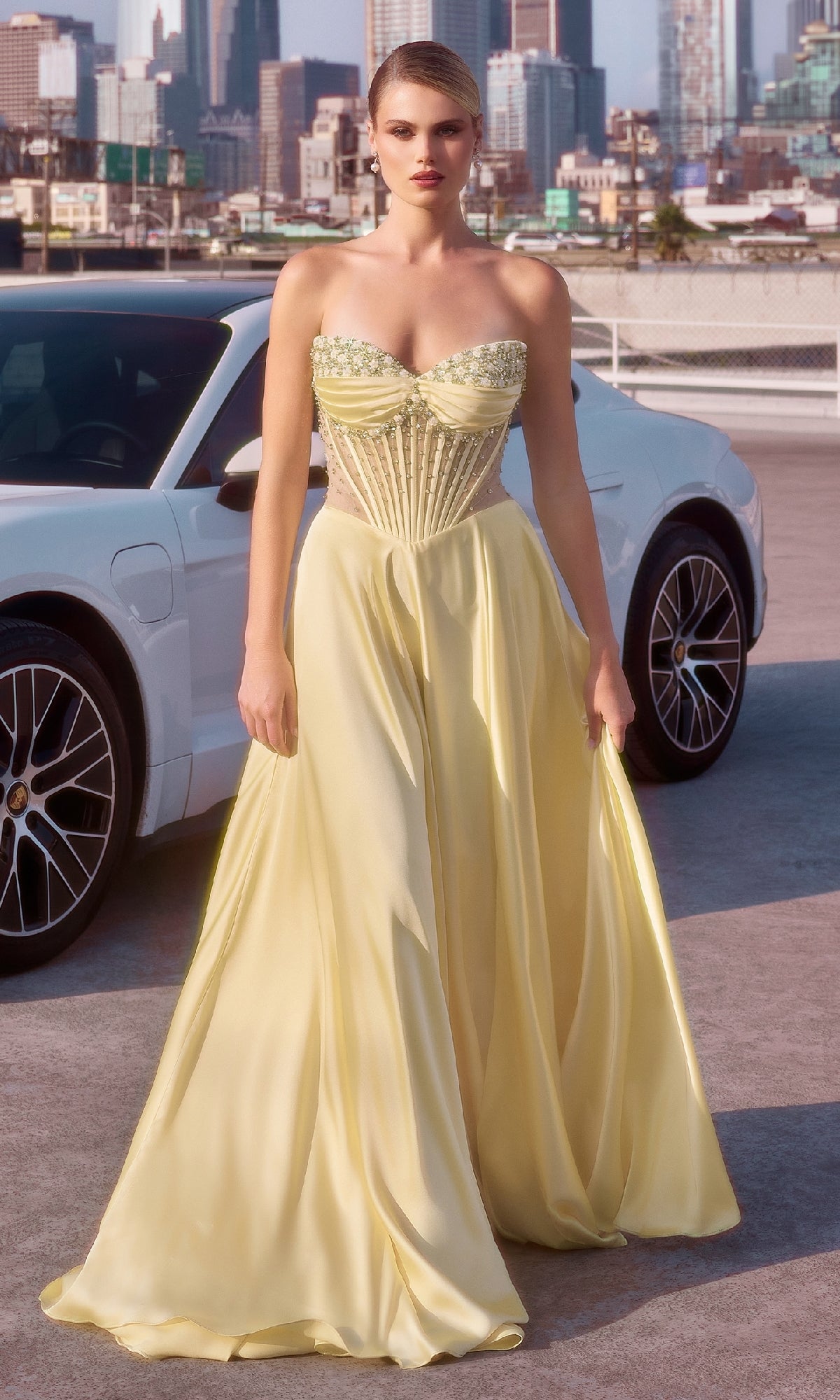 A woman wearing the Ladivine CD297 Strapless Long A-Line Prom Dress—a pale yellow gown with a structured bodice—stands in front of a white car on a city rooftop.